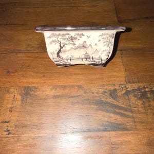 May include: A rectangular ceramic bonsai pot with a cream-colored base and a dark rim. The exterior features a black and white ink-wash style landscape illustration depicting mountains, trees, a bridge, and birds in flight across the surface.