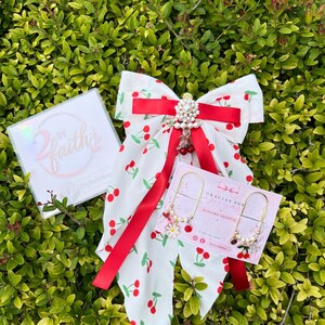 May include: A white hair bow with a red cherry pattern, accented by a red satin ribbon and a central pearl and rhinestone embellishment. Two gold-toned paperclip-style earrings feature small white daisy and red cherry charms with pearls.