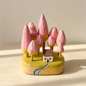 May include: A hand-carved wooden sculpture featuring a small white house with a brown roof nestled among several pink-painted trees with yellow bases. The scene is set on a yellow oval base with a painted blue path leading to the house.