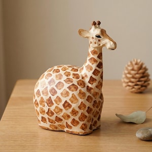 Puede incluir: Una figura de jirafa artesanal con cuerpo crema y manchas marrones. La jirafa tiene un cuello largo y está en posición sentada. La figura está hecha de un material de color claro y se coloca sobre una superficie de madera. Se ve una piña.