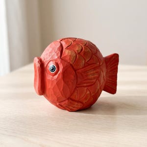 Könnte beinhalten: Eine runde, handgeschnitzte Fischfigur aus Holz, die in einem leuchtenden Orangerot bemalt ist. Die Oberfläche weist sichtbare Meißelspuren und geschnitzte Schuppenmuster auf. Der Fisch hat ein markantes Auge mit schwarzer Pupille und weißer Iris.