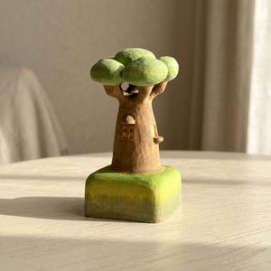 May include: A hand-carved wooden tree sculpture featuring a textured brown trunk with a small window and a rounded green canopy. The tree stands on a square, light green base, showcasing visible chisel marks and a matte finish throughout the piece.
