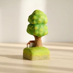 May include: A hand-carved wooden figurine featuring a tall tree with a rounded, textured green canopy and a brown trunk. At the base of the tree sits a small white house with a red roof, all resting on a square, light green grassy platform.