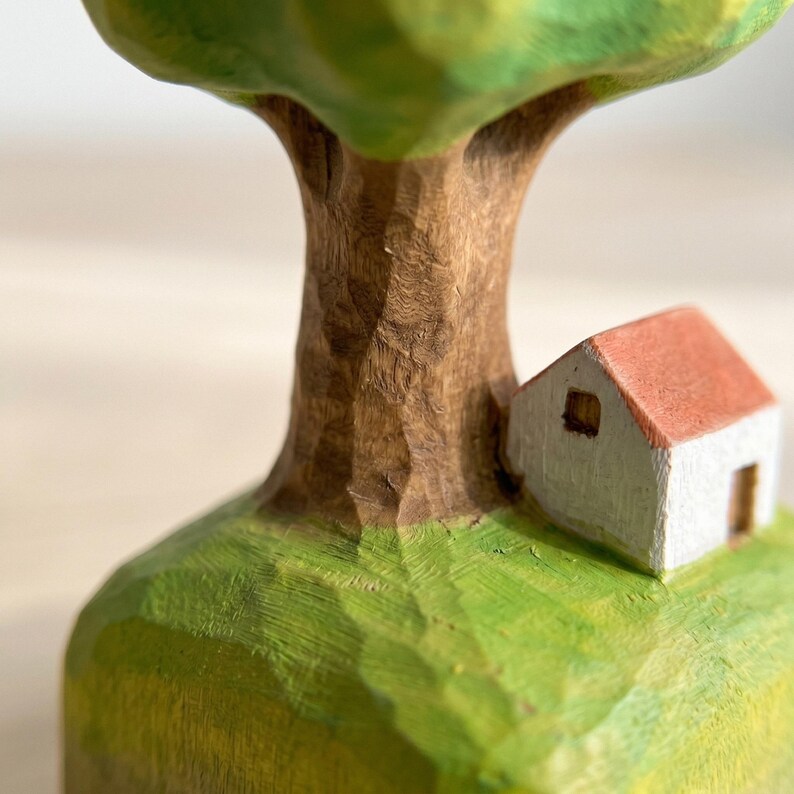 May include: A hand-carved wooden sculpture featuring a green painted tree with a textured brown trunk, standing on a green base next to a small white house with a red roof. The wood grain and chisel marks are visible across the painted surfaces.