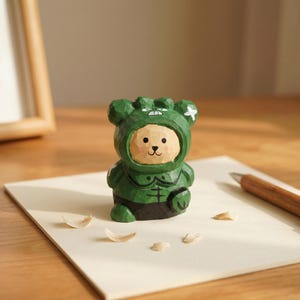 Puede incluir: Figura de madera tallada a mano de un oso con un traje con capucha verde. La cara del oso es de color marr&oacute;n claro con ojos negros y una nariz peque&ntilde;a. El traje tiene una cruz a cada lado de la capucha y un dise&ntilde;o en el pecho. La figura est&aacute; sobre una superficie blanca.