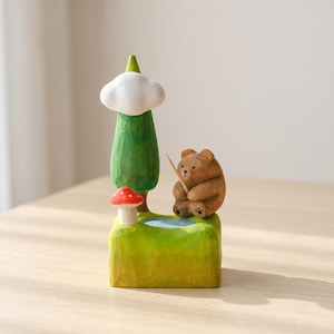 May include: A hand-carved wooden figurine featuring a brown bear sitting by a small blue pond. The scene includes a green tree topped with a white cloud, a red and white mushroom, and a thin wooden stick held by the bear on a grassy base.