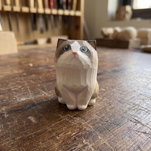 May include: A hand-carved wooden cat figurine featuring a light cream-colored chest and face with brown markings around the ears and eyes. The cat has painted blue eyes, a small pink nose, and visible chisel marks across its smooth wooden surface.