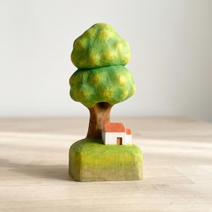 May include: A hand-carved wooden sculpture featuring a tall tree with a rounded, textured green canopy and a brown trunk. At the base of the tree sits a small white house with a red roof, all resting on a square, light green grassy base.