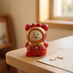 Puede incluir: Figurita de madera tallada a mano de un oso con un atuendo rojo con detalles dorados. El oso tiene una cara sonriente y est&aacute; de pie. La figurita est&aacute; sobre una superficie de madera.