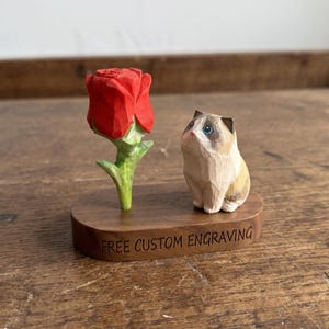 May include: A hand-carved wooden figurine featuring a bright red rose with a green stem next to a small, light brown and white cat with blue eyes. Both are mounted on a dark wood oval base with the text FREE CUSTOM ENGRAVING printed on it.