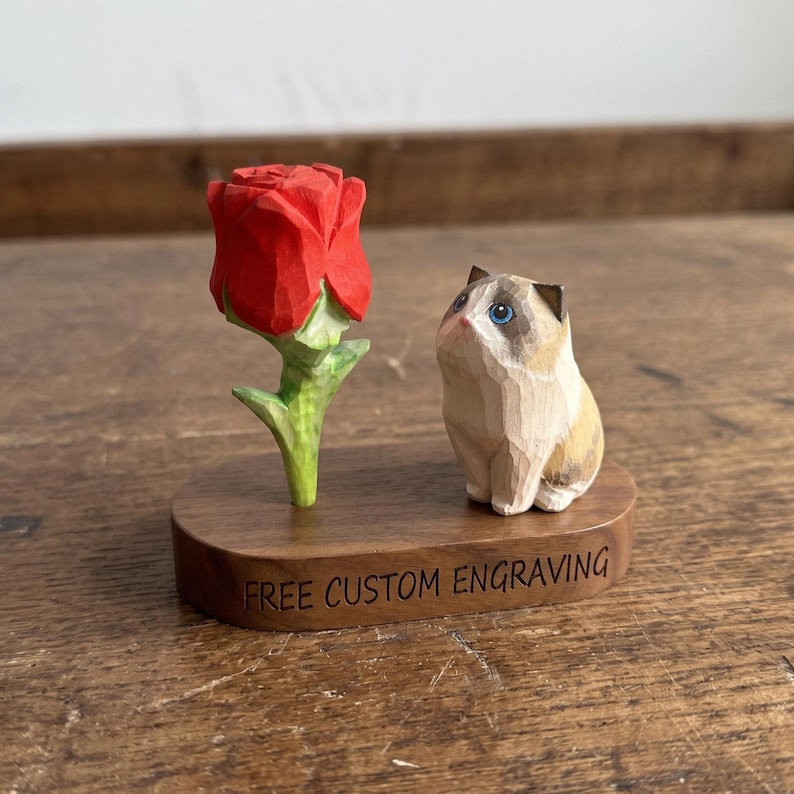 May include: A hand-carved wooden figurine featuring a bright red rose with a green stem next to a small, light brown and white cat with blue eyes. Both are mounted on a dark wood oval base with the text FREE CUSTOM ENGRAVING printed on it.
