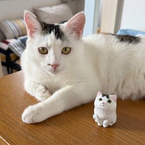 May include: A small, hand-carved wooden figurine of a white cat with black markings on its head and back, featuring painted green eyes and a pink nose, positioned next to a living white cat with similar black markings on its head and back.