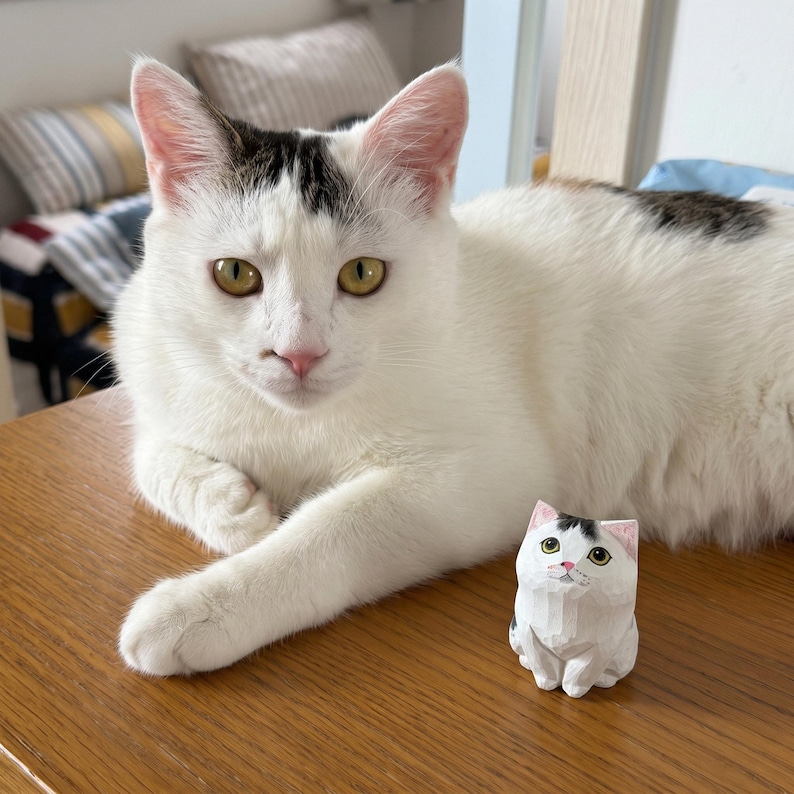 May include: A small, hand-carved wooden figurine of a white cat with black markings on its head and back, featuring painted green eyes and a pink nose, positioned next to a living white cat with similar black markings on its head and back.