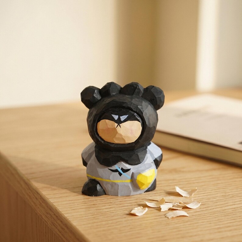 Puede incluir: Una figura de madera tallada de un personaje con forma de oso con un atuendo negro con capucha y un emblema gris y amarillo. La figura tiene una cara de color marr&oacute;n claro y est&aacute; sentada sobre una superficie de madera. Hay virutas de madera esparcidas cerca.