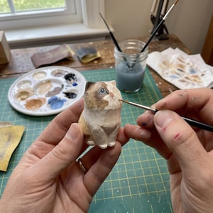May include: A small, hand-carved wooden cat figurine featuring a calico-style pattern with brown, white, and tan painted details. The cat has a pointed face, blue eyes, and a small pink nose, with visible carved facets across its surface.