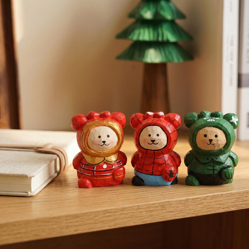 Puede incluir: Tres figuras de osos de madera tallada con disfraces de superh&eacute;roes rojos, rojos y azules y verdes. Los osos tienen caras de color marr&oacute;n claro con ojos de puntos negros y una peque&ntilde;a nariz negra. Las figuras est&aacute;n sobre una superficie de madera.