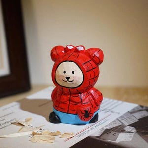 Puede incluir: Una figura de madera tallada de un oso vestido de Spider-Man. El oso es principalmente rojo con detalles de telara&ntilde;a negra, una cara blanca y pantalones cortos azules. Un emblema de ara&ntilde;a negra est&aacute; en el pecho.
