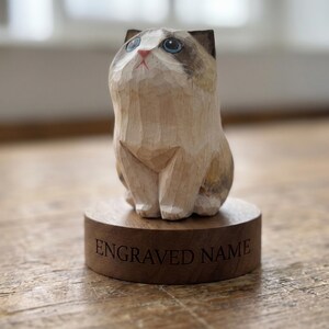 May include: A hand-carved wooden cat figurine with light tan and brown fur markings, blue eyes, and a pink nose. The cat sits on a circular wooden base featuring the text ENGRAVED NAME etched into the front surface in a dark serif font.