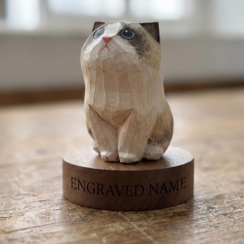 May include: A hand-carved wooden cat figurine with light tan and brown fur markings, blue eyes, and a pink nose. The cat sits on a circular wooden base featuring the text ENGRAVED NAME etched into the front surface in a dark serif font.