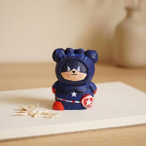 Puede incluir: Una figura de madera tallada de un oso con un disfraz de superh&eacute;roe azul con una estrella blanca y un escudo rojo. El oso tiene una cara sonriente y botas rojas. La figura est&aacute; sobre una superficie blanca.