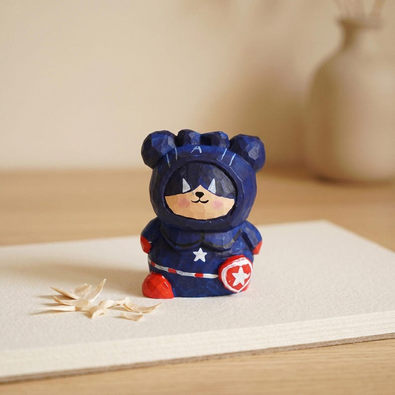 Puede incluir: Una figura de madera tallada de un oso con un disfraz de superh&eacute;roe azul con una estrella blanca y un escudo rojo. El oso tiene una cara sonriente y botas rojas. La figura est&aacute; sobre una superficie blanca.