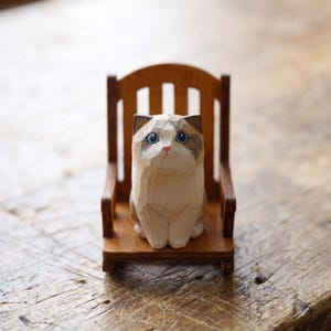 May include: A small, hand-carved wooden figurine of a cat with light cream fur, brown ear markings, and bright blue eyes, sitting upright on a miniature wooden rocking chair with a slatted backrest and armrests, showcasing visible wood grain.