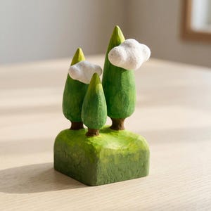 May include: A carved wooden figurine featuring three green trees of varying heights set on a rectangular green base. Two white, fluffy, cloud-like shapes are attached to the sides of the two taller trees, creating a textured, layered landscape.