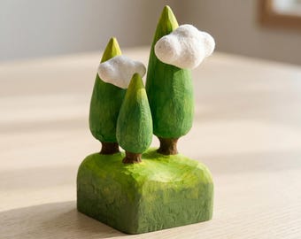 May include: A carved wooden figurine featuring three green trees of varying heights set on a rectangular green base. Two white, fluffy, cloud-like shapes are attached to the sides of the two taller trees, creating a textured, layered landscape.