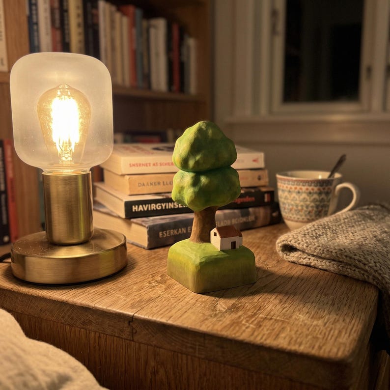 May include: A small, hand-carved wooden sculpture featuring a green tree with a rounded canopy and a tiny white house with a brown roof at its base. The sculpture sits on a square green base, showcasing visible wood grain and painted details.