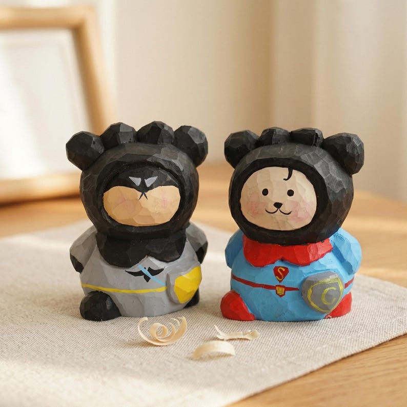 Puede incluir: Dos figuras de oso de madera, una vestida de Batman en gris y amarillo, la otra de Superman en azul y rojo. Ambas tienen capuchas negras con forma de oso y est&aacute;n talladas con vetas de madera visibles.