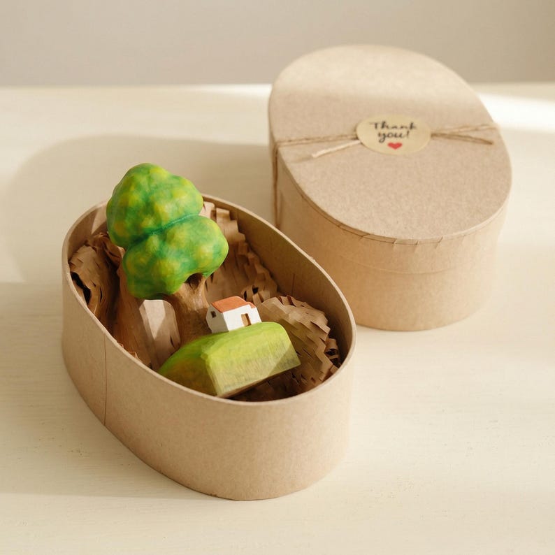 May include: A miniature wooden sculpture of a green tree and a small white house with a red roof, nestled inside an oval cardboard gift box filled with shredded paper. The box lid features a round sticker with the text Thank you and a red heart.