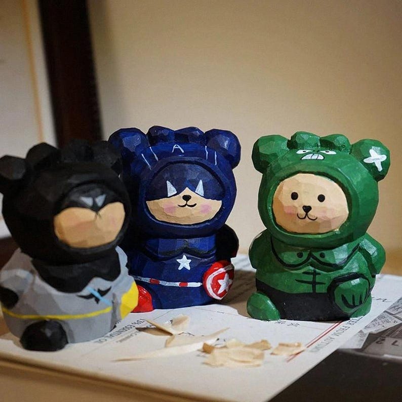 Puede incluir: Tres figuras de osos de madera talladas a mano, disfrazadas de superh&eacute;roes. Los osos est&aacute;n pintados en negro, azul y verde, con detalles como un s&iacute;mbolo de murci&eacute;lago, una estrella y un escudo. Cada oso tiene una expresi&oacute;n facial &uacute;nica.