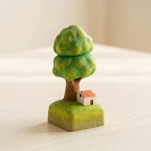 May include: A hand-carved wooden figurine featuring a tree with a rounded, two-tiered green canopy and a brown trunk. At the base of the tree sits a small white house with a terracotta-colored roof, all resting on a square, grassy green base.
