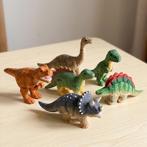 May include: A collection of six hand-carved wooden dinosaur figurines featuring a brown long-necked sauropod, an orange theropod, a green theropod, a green stegosaur with red plates, a green ankylosaur, and a grey triceratops with white spots.