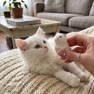 May include: A small, hand-carved wooden figurine of a cat with a smooth, light-colored finish and painted blue eyes. The figurine is held up, positioned near a fluffy white kitten that has one blue eye and one green eye, looking directly at the object.