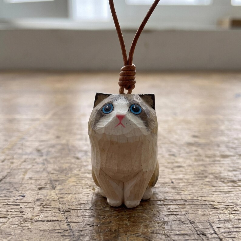 May include: A hand-carved wooden cat pendant featuring a light tan and cream coat with dark brown ear markings. The cat has bright blue eyes and a small pink nose, suspended from a thin brown leather cord tied in a knot at the top of the head.