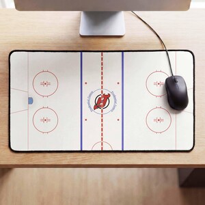 May include: A rectangular desk mat featuring a white ice hockey rink graphic with red and blue lines, face-off circles, and the New Jersey Devils logo at center ice with the text Prudential Center. A black computer mouse sits on the right side.