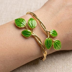 May include: A gold-toned metal cuff bracelet featuring a twisted vine design adorned with vibrant green enameled leaves. The bracelet wraps around the wrist, showcasing two distinct branches that terminate in clusters of detailed green leaves.