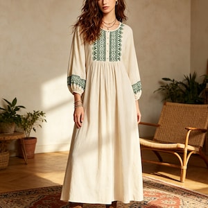 Puede incluir: Un vestido largo de color crema con mangas largas, que presenta bordados de color verde oscuro alrededor del cuello redondo, en el centro del pecho y en los puños. El vestido tiene una cintura fruncida y una falda fluida, combinado con botas marrones.