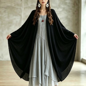 Könnte beinhalten: Ein hellgraues ärmelloses Kleid mit einem asymmetrischen Lagenrock, kombiniert mit einer langen, vorne offenen schwarzen Strickjacke. Das Outfit umfasst eine Halskette mit einem kleinen roten Anhänger, weiße Ohrhänger und braune Riemchensandalen.