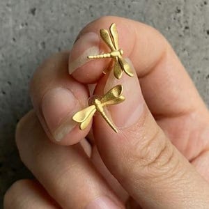 May include: A pair of small, gold-toned dragonfly stud earrings with a matte finish. Each earring features a detailed insect body with delicate, outstretched wings, designed to be worn as a matching set on the earlobes for a subtle accessory.
