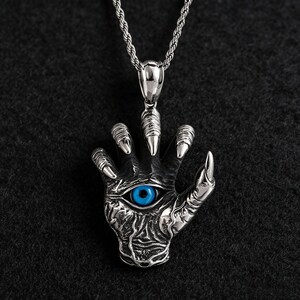 Könnte beinhalten: Silberfarbene Halskette mit einem Anhänger in Form einer Hand mit scharfen Krallen und einem blauen Auge. Die Kette hängt an einer silbernen Seilkette und hat ein gotisches oder Fantasy-Thema.