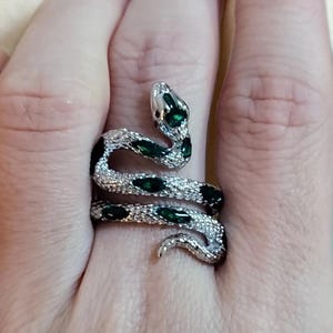 May include: A silver-toned snake ring featuring a textured, scale-like surface and inlaid teardrop-shaped dark green stones along the body. The snake wraps around the finger, with the head positioned at the top and the tail curving downward.