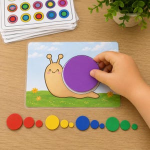 Op de afbeelding: Een educatieve set bestaande uit een rechthoekige kaart met een slakillustratie en een paars cirkelvormig onderdeel. De set bevat een stapel voorbeeldkaarten en diverse kleurrijke cirkelvormige schijven in rood, geel, blauw en groen.