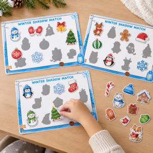 May include: Three rectangular Winter Shadow Match activity boards featuring colorful holiday-themed stickers like snowmen, Christmas trees, and snowflakes. Each board displays gray silhouettes for matching, with loose stickers of a candy cane, mittens, and animals nearby.