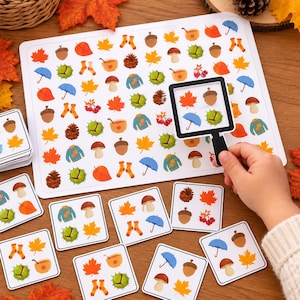 Könnte beinhalten: Ein Zuordnungsspiel mit einem weißen Spielbrett und quadratischen Karten, bedruckt mit herbstlichen Symbolen wie orangefarbenen Ahornblättern, braunen Eicheln, blauen Regenschirmen, Tannenzapfen, Pilzen, grünen Kastanien, orangefarbenen Socken und Pullovern.