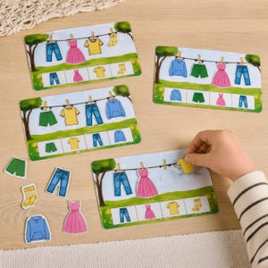 Peut inclure: Un ensemble de cartes éducatives illustrant des vêtements tels que des jeans bleus, des robes roses, des t-shirts jaunes, des shorts verts et des chaussettes jaunes suspendus à une corde à linge, avec des icônes correspondantes en bas.