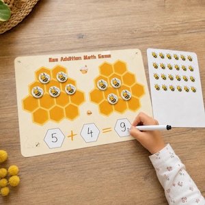 Könnte beinhalten: Ein Bee Addition Math Game mit zwei gelben Wabenmustern, kleinen Bienen-Spielsteinen und einem weißen Bogen mit Bienenaufklebern. Das Spielbrett zeigt den Text Bee Addition Math Game sowie eine mathematische Gleichung mit den Zahlen 5, 4 und 9.