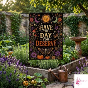 May include: A rectangular garden flag featuring a black background with a central sun, crescent moons, stars, butterflies, mushrooms, pumpkins, and flowers. The text reads HAVE THE DAY YOU DESERVE in gold lettering. The flag is displayed on a metal stand.
