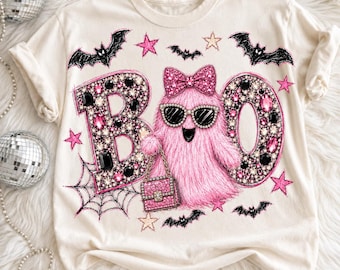 Può includere: Una t-shirt color crema con la grafica di un fantasma rosa peloso che indossa un fiocco rosa e occhiali da sole neri. Il fantasma tiene una borsa rosa. Le lettere B e O sono riempite di gemme rosa e nere. Pipistrelli e stelle lo circondano.